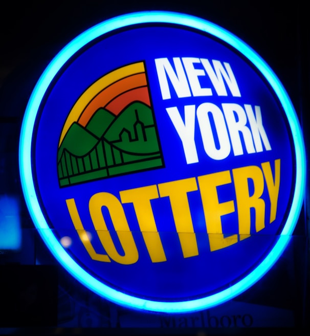 A bright, circular neon sign with a blue background, featuring the words 'New York Lottery' in bold white and yellow letters. The sign includes a colorful illustration of a landscape with green mountains, an orange-red sunset, and a bridge, all encapsulated within the circle.