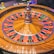 A roulette wheel with alternating red and black numbered pockets on a wooden platform. The wheel contains a small white ball resting in one of the pockets, indicating gameplay. Surrounding the wheel are stacks of colorful poker chips, suggesting a casino environment.