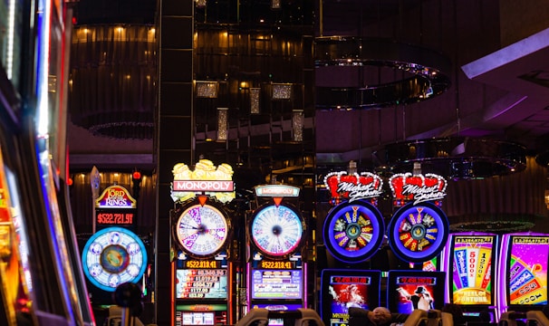 A lively casino scene featuring a row of bright, colorful slot machines with themes like Monopoly, Michael Jackson, and Lord of the Rings. The gaming machines display a variety of flashing lights and digital screens, creating an energetic and exciting atmosphere. Reflections of the lights are visible in glossy surfaces above the machines.