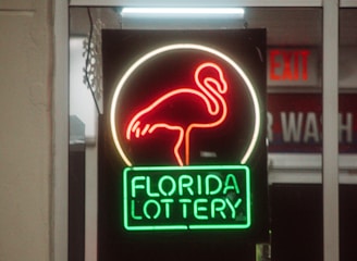 A neon sign depicting a flamingo above the words 'Florida Lottery' is prominently displayed. The sign is illuminated in bright red and green colors, and it is framed by glass panels. An 'EXIT' sign is visible in the background.