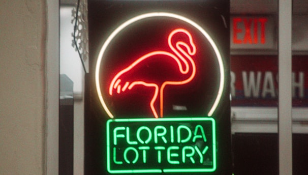 A neon sign depicting a flamingo above the words 'Florida Lottery' is prominently displayed. The sign is illuminated in bright red and green colors, and it is framed by glass panels. An 'EXIT' sign is visible in the background.
